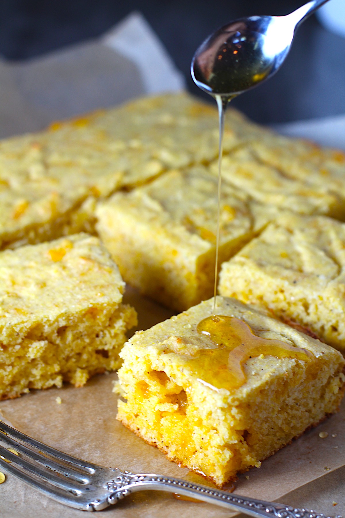 Cheddar Honey Glazed Cornbread pieces cut on parchment paper with honey being drizzled on top and fork and crumbs around.