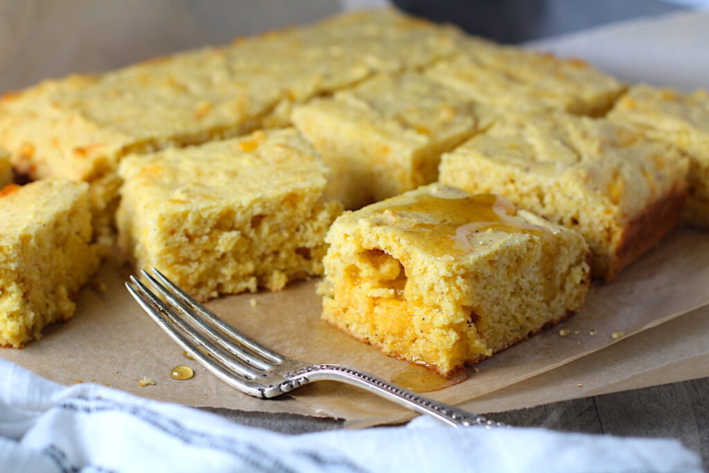 Cheddar Honey Gluten Free Cornbread pieces stacked on parchment paper with honey drizzled on top and fork and crumbs around.