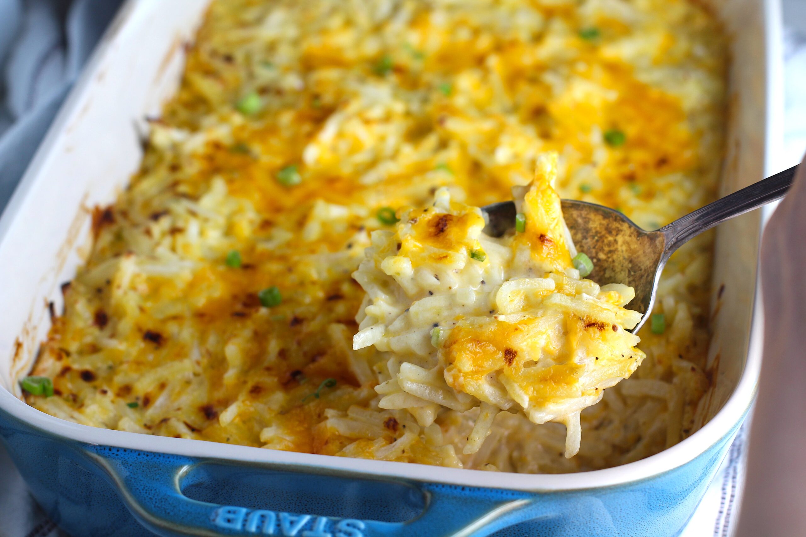 Golden brown cheesy Hash Brown Casserole without Soup in a Staub Casserole dish on counter with a rustic serving spoon scooping up a cheesy spoonful.