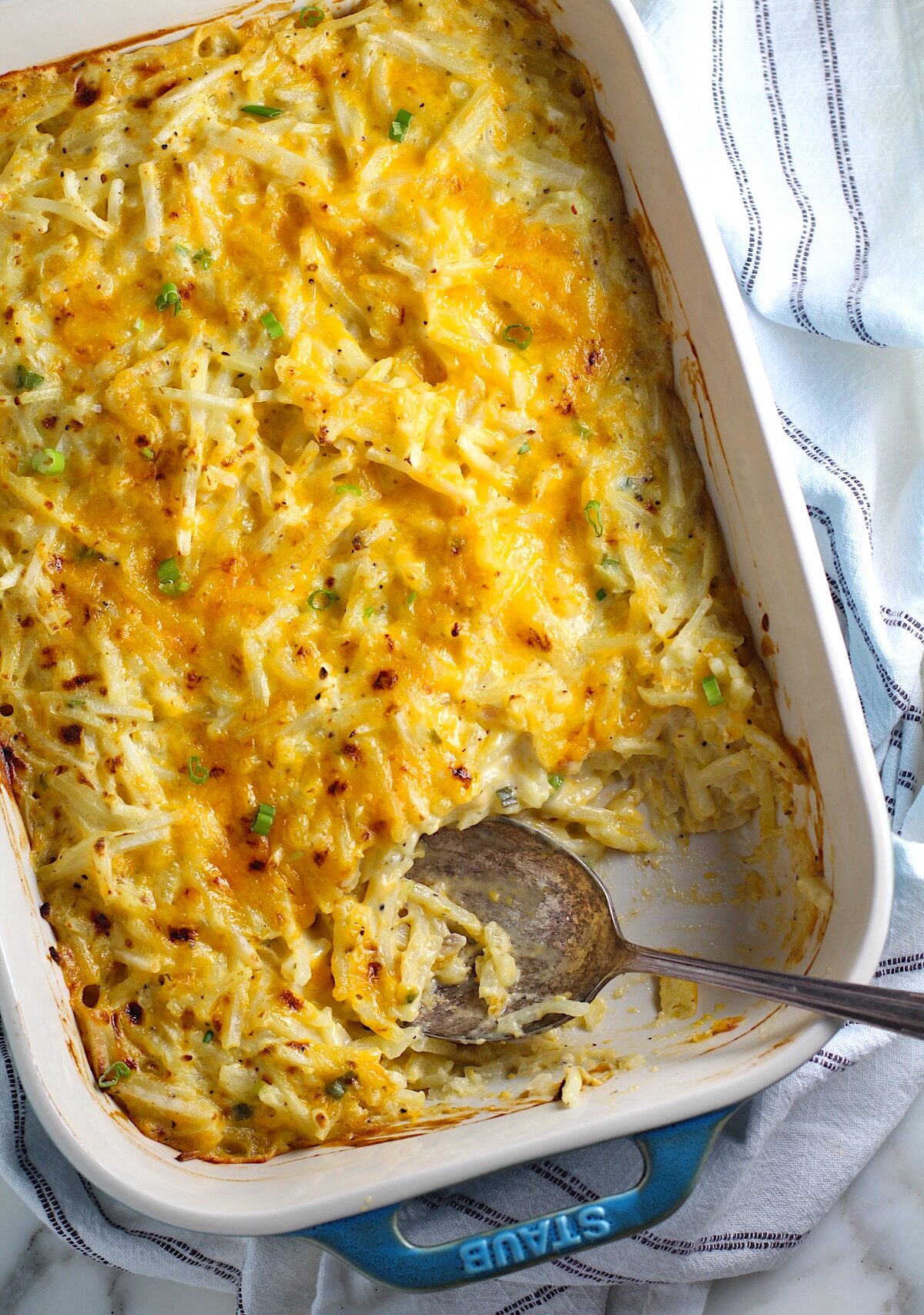 Golden brown cheesy Hash Brown Casserole without Soup in a Staub Casserole dish on counter with a rustic serving spoon in the corner where some casserole has been taken out.
