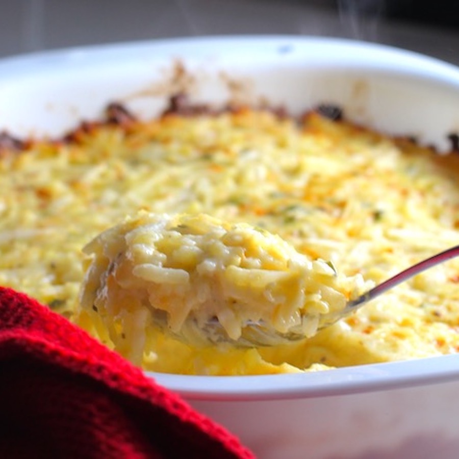 Spoon scooping Cheesy Hash Brown Casserole in casserole dish on counter with wine glass in background. This recipe does not disappoint! It's warm, creamy, and full of flavor! And it's the perfect side dish for your holiday dinner.