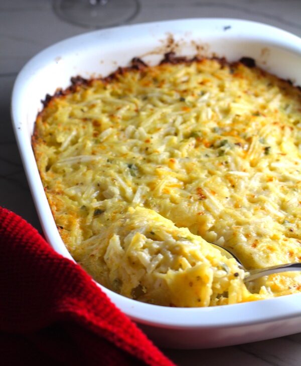 This is a photo of Cheesy Hash Brown Casserole in a white casserole dish with a serving spoon, ready to serve.