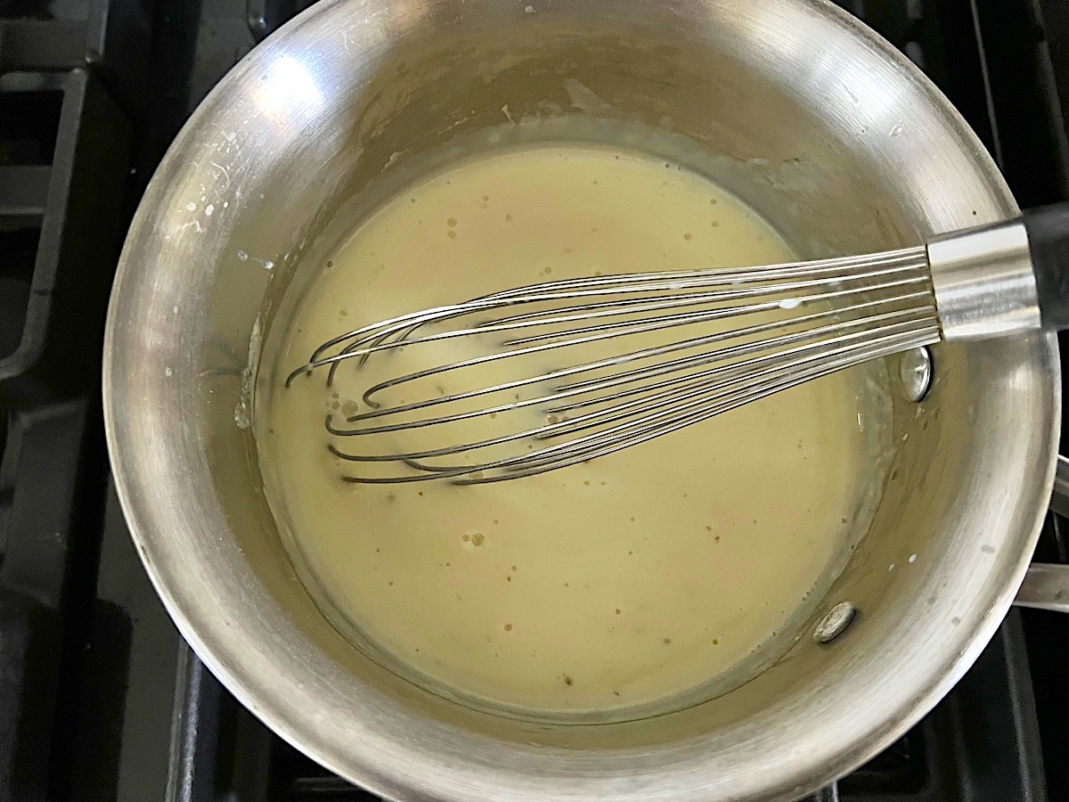 Flour, butter, milk, broth being whisked in a saucepan on the stove for Hash Brown Casserole without Soup.