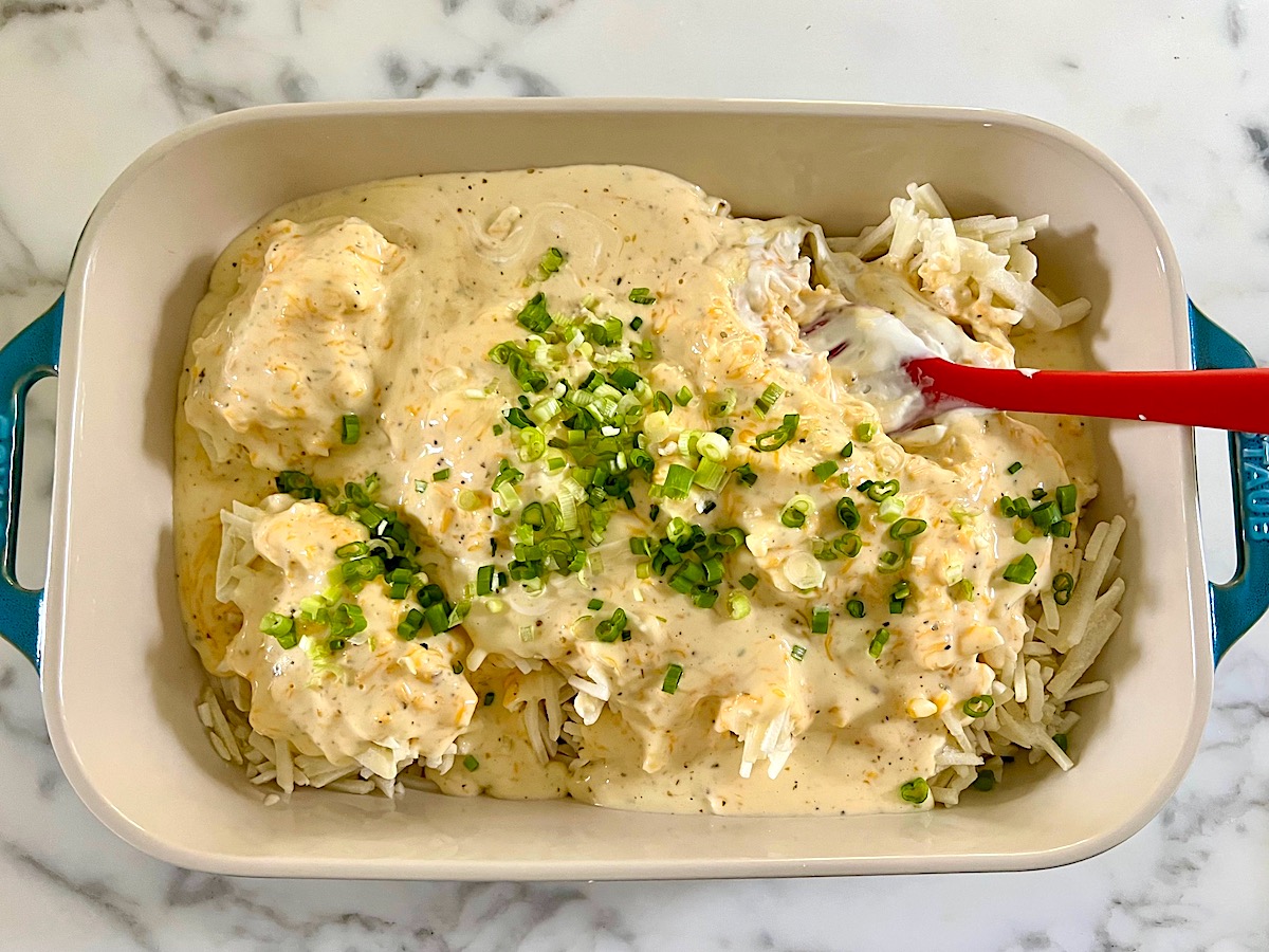 Cheese sauce and scallions added on top of the frozen hash browns in a casserole dish on counter before being mixed for Hash Brown Casserole without Soup.