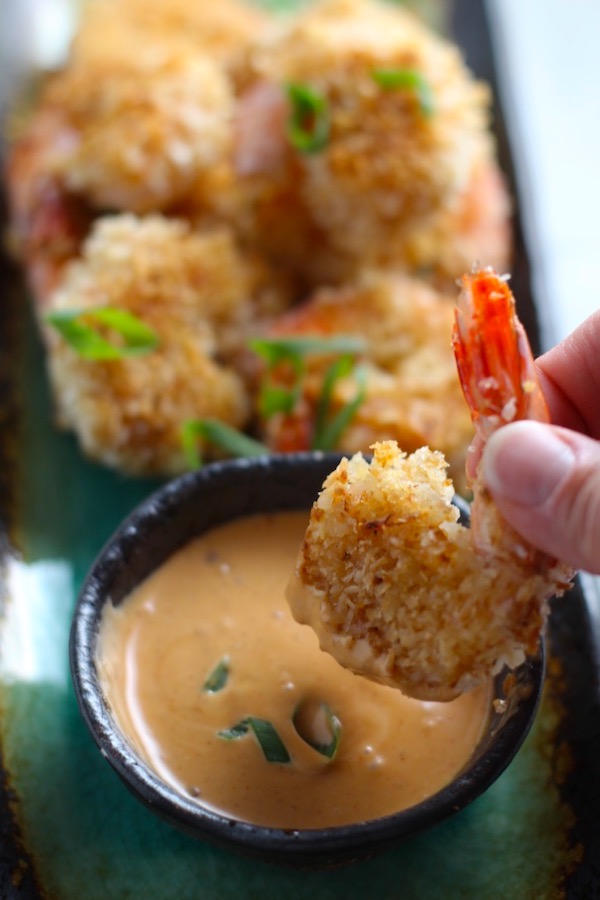 Baked Coconut Shrimp on plate with hand dipping one into Sriracha Sauce. Crunchy and utterly irresistible, this 30-Minute Baked Coconut Shrimp recipe is one that you have got to try!  They are baked, not fried, so they are EASY to cook and HEALTHIER for you too! 