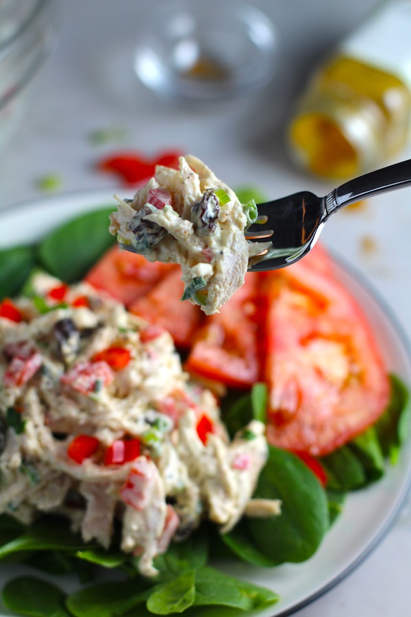 Fork with bite of Curry Chicken Salad on spinach with tomato slices. It has so many fantastic flavors and textures!  The chicken breast is simply roasted and mixed with a creamy sweet and savory curry dressing.  Red pepper gives you a fresh crunch, scallion gives a savory bite, and raisins give a burst of sweet.  #chicken #chickenrecipes #chickensalad #currychicken #mealprep #lunch #healthyrecipes