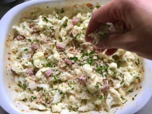 Hand sprinkling topping on Cauliflower Cordon Bleu Bake recipe. It layers in nutty and salty Gruyere Cheese, with a creamy Vegetable broth based sauce with garlic and parsley.  Then the ham and cauliflower are folded into all of this goodness and it's baked in the oven until it all becomes one knock-your-socks-off dish! #cauliflower #lowcarb #glutenfree #casserole #onepot #familydinner #mealprep #easydinner #dinner #healthydinner #healthyfood #healthyrecipes