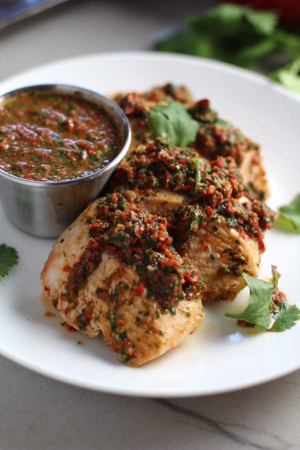 Chicken Chimichurri Rojo on a plate with fresh cilantro and a cup of chimichurri. It is roasted Chicken topped with a cool, fresh, tangy, and incredibly full-flavored!  The Rojo comes from the addition of red pepper.  All of the ingredients simply get blended in a food processor so it could not be easier!  #chicken #chickenrecipes #chimichurri #easychicken #easydinners #dinner