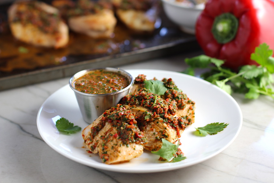 Chicken Chimichurri Rojo on a plate with cup of chimichurri. It is broiled or grilled Chicken topped with a cool, fresh, tangy, and incredibly full-flavored! The Rojo comes from the addition of red pepper. All of the ingredients simply get blended in a food processor so it could not be easier! #chicken #chickenrecipes #chimichurri #easychicken #easydinners #dinner