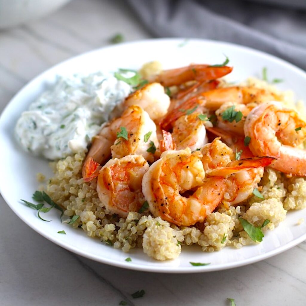 Garlic Lime Grilled Greek Shrimp with Tzatziki Sauce over Quinoa on plate. The Shrimp is simply sauteed with minced garlic, olive oil, and lime for big bold flavors. The Tzatziki Sauce is bright and cool with creamy yogurt with fresh cucumber, lime, garlic, and scallion.