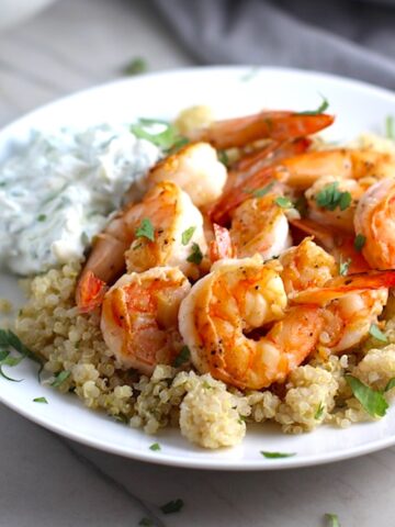 Garlic Lime Grilled Greek Shrimp with Tzatziki Sauce over Quinoa on plate. The Shrimp is simply sauteed with minced garlic, olive oil, and lime for big bold flavors. The Tzatziki Sauce is bright and cool with creamy yogurt with fresh cucumber, lime, garlic, and scallion.