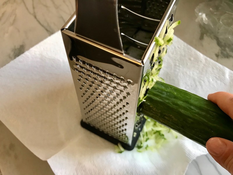 Shredding cucumber on grater over paper towel for Garlic and Lime Shrimp with Tzatziki Sauce. The Shrimp is simply sauteed with minced garlic, olive oil, and lemon for big bold flavors. The Tzatziki Sauce is bright and cool with creamy yogurt with fresh cucumber, lime, garlic, and scallion. #shrimp #easydinner #dinner #seafoodrecipes #shrimprecipes