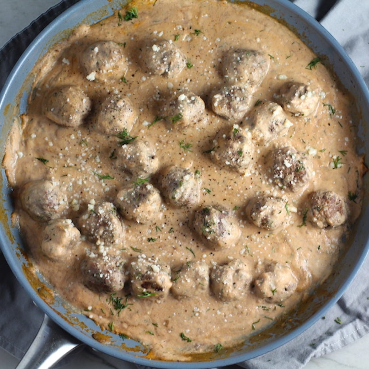 Meatballs in Cauliflower Cheese Sauce in skillet on towel on counter with fresh dill on the side. The Meatballs are in a creamy cauliflower sauce with garlic, fresh dill, cream cheese, and tomato paste.