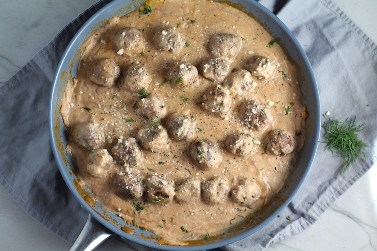 Meatballs in Cauliflower Cheese Sauce in skillet on towel on counter with fresh dill on the side. The Meatballs are in a creamy cauliflower sauce with garlic, fresh dill, cream cheese, and tomato paste.