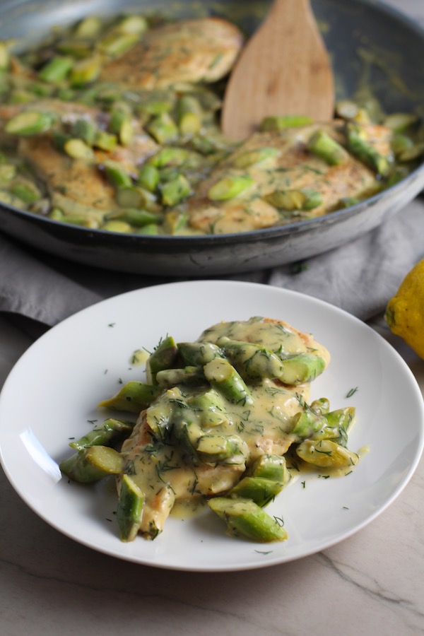 Lemon Dill Sauce Chicken and Asparagus on plate with pan in back. It has chicken in a light and creamy sauce flavored with garlic, lemon and fresh dill. It's loaded with fresh asparagus. #springrecipes #dinner #easydinner #healthydinner #lemon #asparagus #chicken #glutenfree #easyrecipes