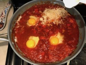 Shredded cheese being sprinkled over raw eggs sitting in tomato sauce in pan for Skillet Eggs in Spicy Tomato Pepper Sauce with Manchego Cheese. With a smoky, spicy, thick, and luscious red pepper tomato sauce. #eggs #huevosrancheros #tomatosauce #easydinner #brunch #shakshuka