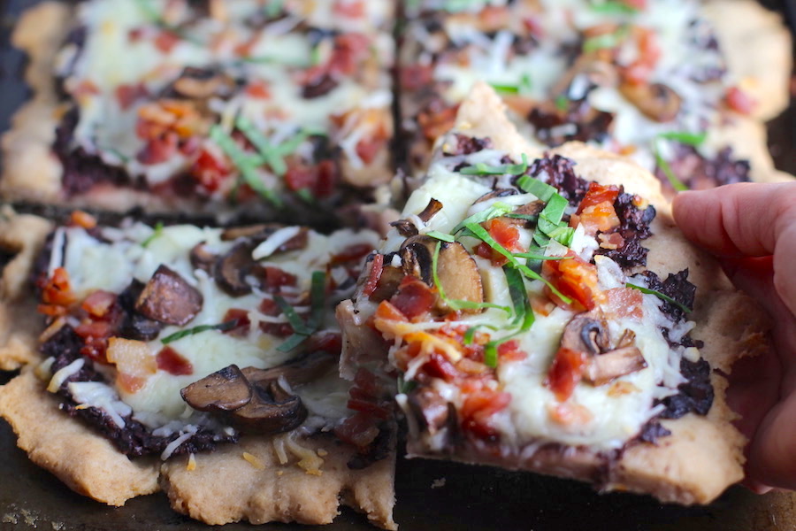 Hand holding a piece of Flatbread cut into 4 pieces on a pan with a crispy gluten free crust, salty and briny Olive Tapenade, creamy mozzarella, earthy and meaty mushrooms, peppery pancetta, and fresh basil. #flatbread #pizza #olives #tapenade #mediterranean #easydinner #dinner