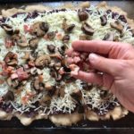 Hand putting pancetta and mushrooms on cheese on flatbread for Olive & Pancetta Flatbread with mozzarella, mushrooms, and fresh basil. #flatbread #pizza #olives #tapenade #mediterranean #easydinner #dinner