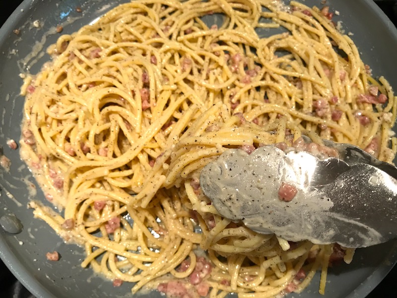 Tongs mixing Pancetta, Parmesan, and Pepper Pasta in a skillet. The Pancetta gives a salty and slightly peppery flavor, Parmesan cheese creates a nutty and creamy sauce, and the ground black pepper gives a peppery flavor that makes this pasta stand out. #pasta #easypasta #easydinner #dinner #italian #familydinner #onpotdinners #onepandinners #parmesan