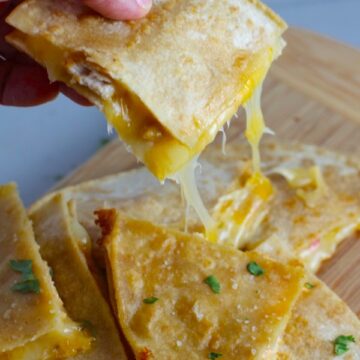 Hand holding Chicken Corn Tortilla Quesadilla triangle with melty cheese pulling. They have crispy edges, a soft gooey center, and are mouthwatering flavor. They have hearty shredded chicken flavored with warm and smoky Mexican spices. Then shredded Pepper Jack cheese and Cheddar is layered to get melty and oozy and delicious. It's an irresistible new easy dinner or appetizer idea.