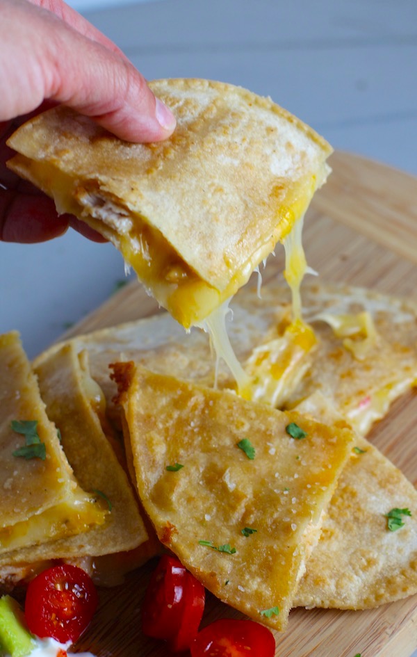 Hand holding Chicken Corn Tortilla Quesadilla triangle with melty cheese pulling. They have crispy edges, a soft gooey center, and are mouthwatering flavor. They have hearty shredded chicken flavored with warm and smoky Mexican spices. Then shredded Pepper Jack cheese and Cheddar is layered to get melty and oozy and delicious. It's an irresistible new easy dinner or appetizer idea. #quesadillas #mexicanfood #enchiladas #familydinner #chicken