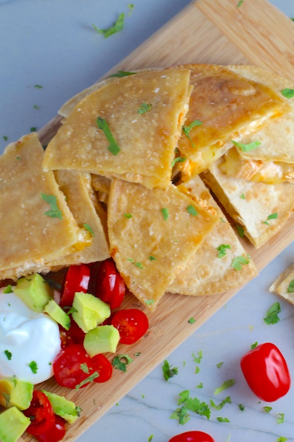 Chicken Corn Tortilla Quesadilla triangles on platter with avocado, tomato, & sour cream.  They have crispy edges, a soft gooey center, and are mouthwatering flavor.  They have hearty shredded chicken flavored with warm and smoky Mexican spices.  Then shredded Pepper Jack cheese and Cheddar is layered to get melty and oozy and delicious.  It's an irresistible new easy dinner or appetizer idea.  #quesadillas #mexicanfood #enchiladas #familydinner #chicken