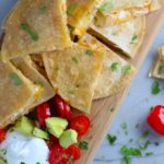 Chicken Corn Tortilla Quesadilla triangles on platter with avocado, tomato, & sour cream. They have crispy edges, a soft gooey center, and are mouthwatering flavor. They have hearty shredded chicken flavored with warm and smoky Mexican spices. Then shredded Pepper Jack cheese and Cheddar is layered to get melty and oozy and delicious. It's an irresistible new easy dinner or appetizer idea. #quesadillas #mexicanfood #enchiladas #familydinner #chicken