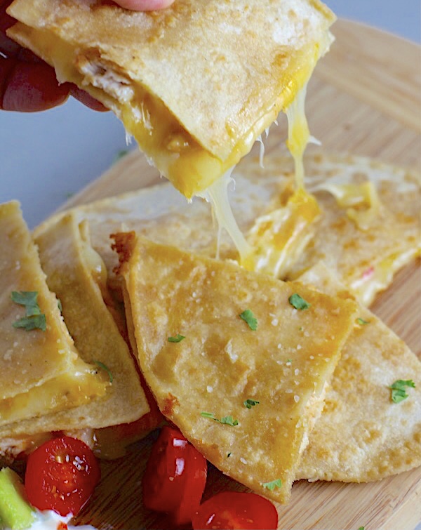 Hand holding Chicken Corn Tortilla Quesadilla triangle with melty cheese pulling. They have crispy edges, a soft gooey center, and are mouthwatering flavor. They have hearty shredded chicken flavored with warm and smoky Mexican spices. Then shredded Pepper Jack cheese and Cheddar is layered to get melty and oozy and delicious. It's an irresistible new easy dinner or appetizer idea.