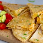 Chicken Corn Tortilla Quesadilla triangles on platter with avocado, tomato, & sour cream. They have crispy edges, a soft gooey center, and are mouthwatering flavor. They have hearty shredded chicken flavored with warm and smoky Mexican spices. Then shredded Pepper Jack cheese and Cheddar is layered to get melty and oozy and delicious. It's an irresistible new easy dinner or appetizer idea. #quesadillas #mexicanfood #enchiladas #familydinner #chicken