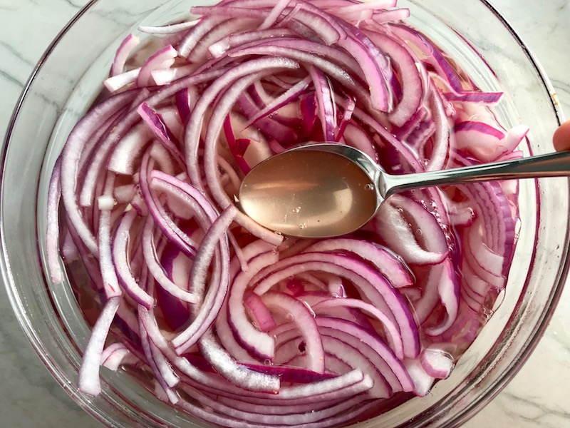 Slices of red onion with pickling liquid in bowl for Pickled Onions in a bowl soaking. They are tart, tangy, sweet, and savory.  They go on just about anything and are so unbelievably delicious, they can make a shoe taste good!  Best part, you can make a ton of them ahead!