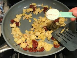 Adding flour to chopped artichoke hearts and sun dried tomatoes in pan for Skillet Artichoke Chicken with sun dried tomatoes, basil, garlic, in cream sauce. #skilletchicken #skilletdinner #chicken #easydinner #familydinner #italian #tuscanchicken
