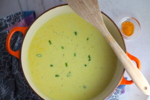 Chilled Golden Corn Soup in a pot with a wooden spoon on side and turmeric on counter. Scallions on top for garnish. It's thick, creamy, silky and delicious.  The entire family will love this easy stovetop corn soup! #summerfood #cornsoup #corn #vegetarian #vegetablerecipes #sides