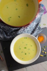 Chilled Golden Corn Soup in a bowl with pot of soup behind and turmeric spice in a bowl on the side. Scallions on top for garnish. It's thick, creamy, silky and delicious.  The entire family will love this easy stovetop corn soup! #summerfood #cornsoup #corn #vegetarian #vegetablerecipes #sides