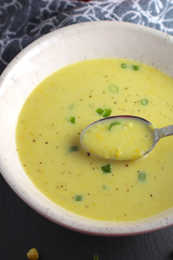 Closeup of Chilled Golden Corn Soup in a bowl with a spoon in it. Scallions on top for garnish. It's thick, creamy, silky and delicious.  The entire family will love this easy stovetop corn soup! #summerfood #cornsoup #corn #vegetarian #vegetablerecipes #sides
