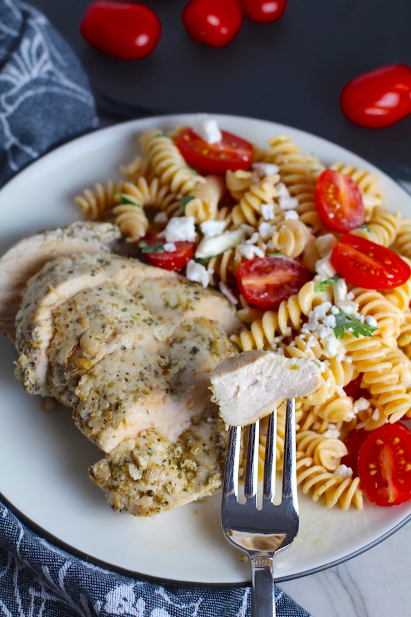 Fork with bite of Mediterranean Chicken sliced on plate with a tomato and feta pasta. This Mediterranean Marinade is only a few ingredients, can be made ahead, and is delicious!