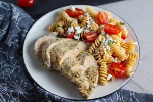 Mediterranean Chicken sliced on plate with a tomato and feta pasta. This Mediterranean Marinade is only a few ingredients, can be made ahead, and is delicious!
