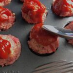 Spoon spreading ketchuo on Raw Mini Meatloaf Muffins in muffin tin. They have only 5 simple ingredients, cook in 30 minutes, and are gluten free.