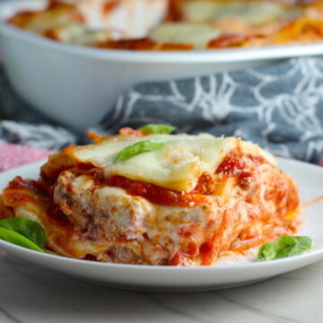 Ground Chicken Layered Lasagna Recipe on a plate with fresh basil with casserole dish in background. from the dish. This Italian Sausage-style chicken Lasagne Recipe is the stuff that dreams are made of.  Layers of pasta, creamy ricotta, salty chewy mozzarella, salty and hearty Italian Sausage, and sweet and tangy tomato sauce.