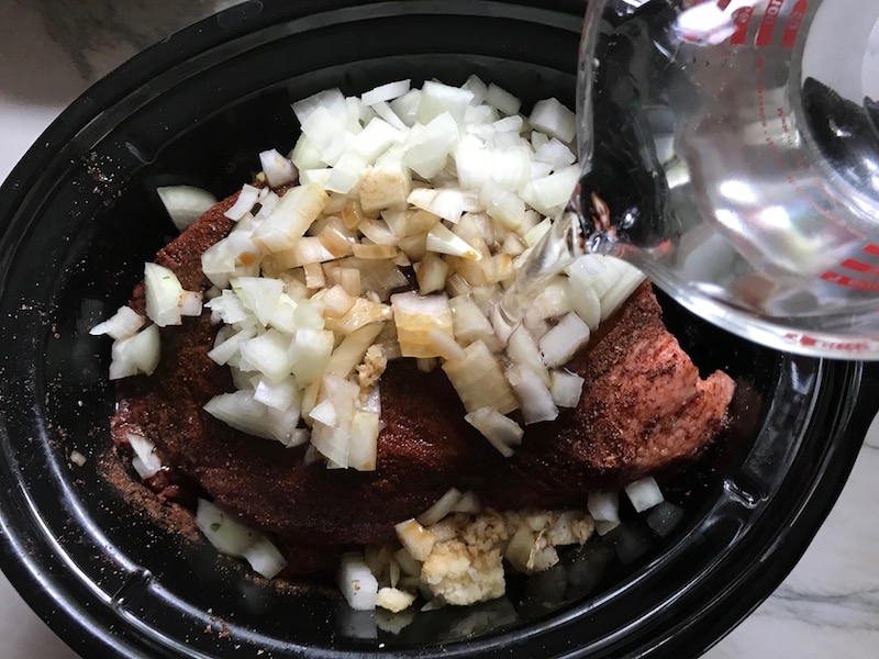 Adding water to onions and raw brisket in Slow Cooker insert for Smoky Pulled Beef Brisket.  It's smokey, savory, tender, moist, and full of flavor. In a delicious sauce, it goes great on a sandwich, over polenta, on rice, or potatoes. #slowcooker #crockpot #brisket #beefbrisket #pulledbeef #pulledpork