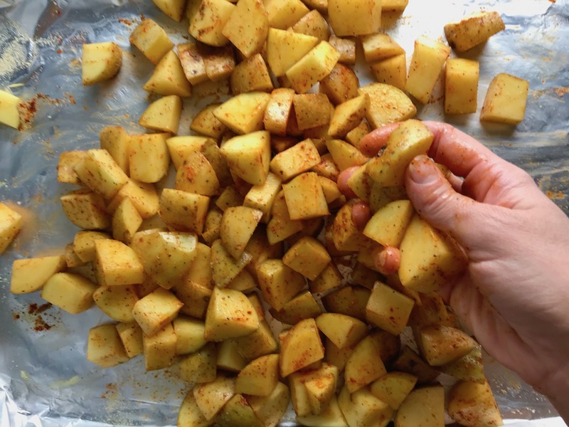 Hand rubbing seasoning on cut potatoes on sheet pan for Patatas Bravas with chorizo and creamy Paprika aioli.  This Patatas Bravas Recipe, or Spicy Potatoes, is easy, indulgent, and utterly delicious!  #potatorecipes #potatoes #potatosidedishes #patatasbravas #skilletpotatoes #sidedishes #chorizo