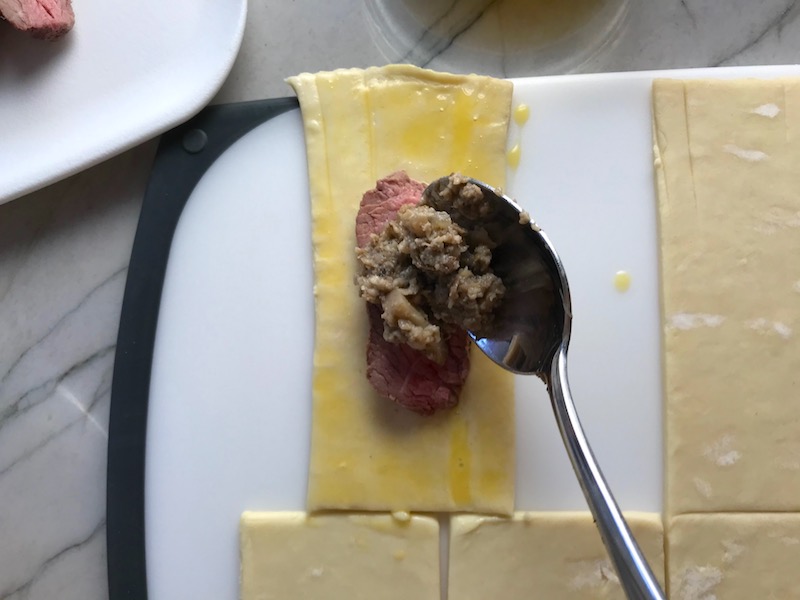 Spoon adding mushroom filling on top of slice of beef on cut Puff Pastry for Mini Beef Wellington Bites. They are flaky and buttery Puff Pastry is filled with a creamy mushroom and parmesan filling and tender beef filet.  #appetizers #partyfood