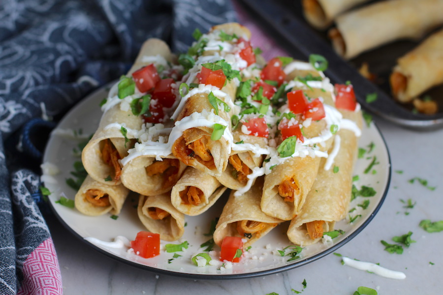Baked Chicken Taquitos stacked on plate with toppings.  Corn tortillas are filled with smoky taco seasoned shredded chicken, and creamy cheddar cheese.  #dinnerideas #familydinner #chickenrecipes #chickendinner #tacos #taquitos