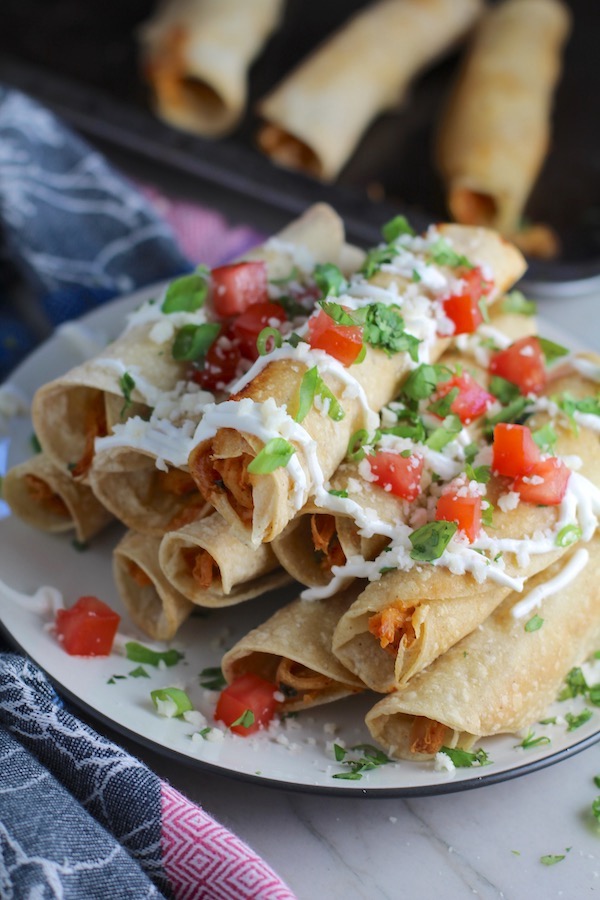 Baked Chicken Taquitos stacked on plate with toppings. Corn tortillas are filled with smoky taco seasoned shredded chicken, and creamy cheddar cheese.  #dinnerideas #familydinner #chickenrecipes #chickendinner #tacos #taquitos