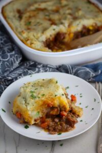 Piece of Cottage Pie on plate with dish in back. This recipe has Spanish Chorizo and ground beef cooked in a rich and savory gravy with veggies and herbs.  Creamy mashed potatoes sit on top with manchego, parmesan, and garlic. #dinnerideas #cottagepie #shepherdspie
