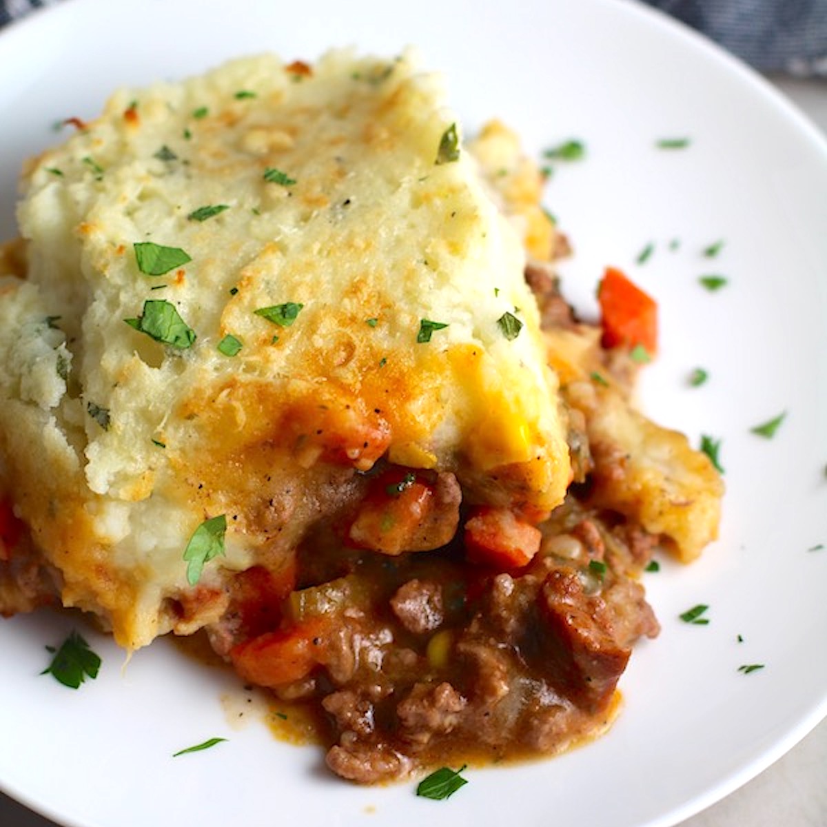Piece of Sausage Shepherds Pie on plate with dish in back. This recipe has Spanish Chorizo and ground beef cooked in a rich and savory gravy with veggies and herbs.  Creamy mashed potatoes sit on top with manchego, parmesan, and garlic.