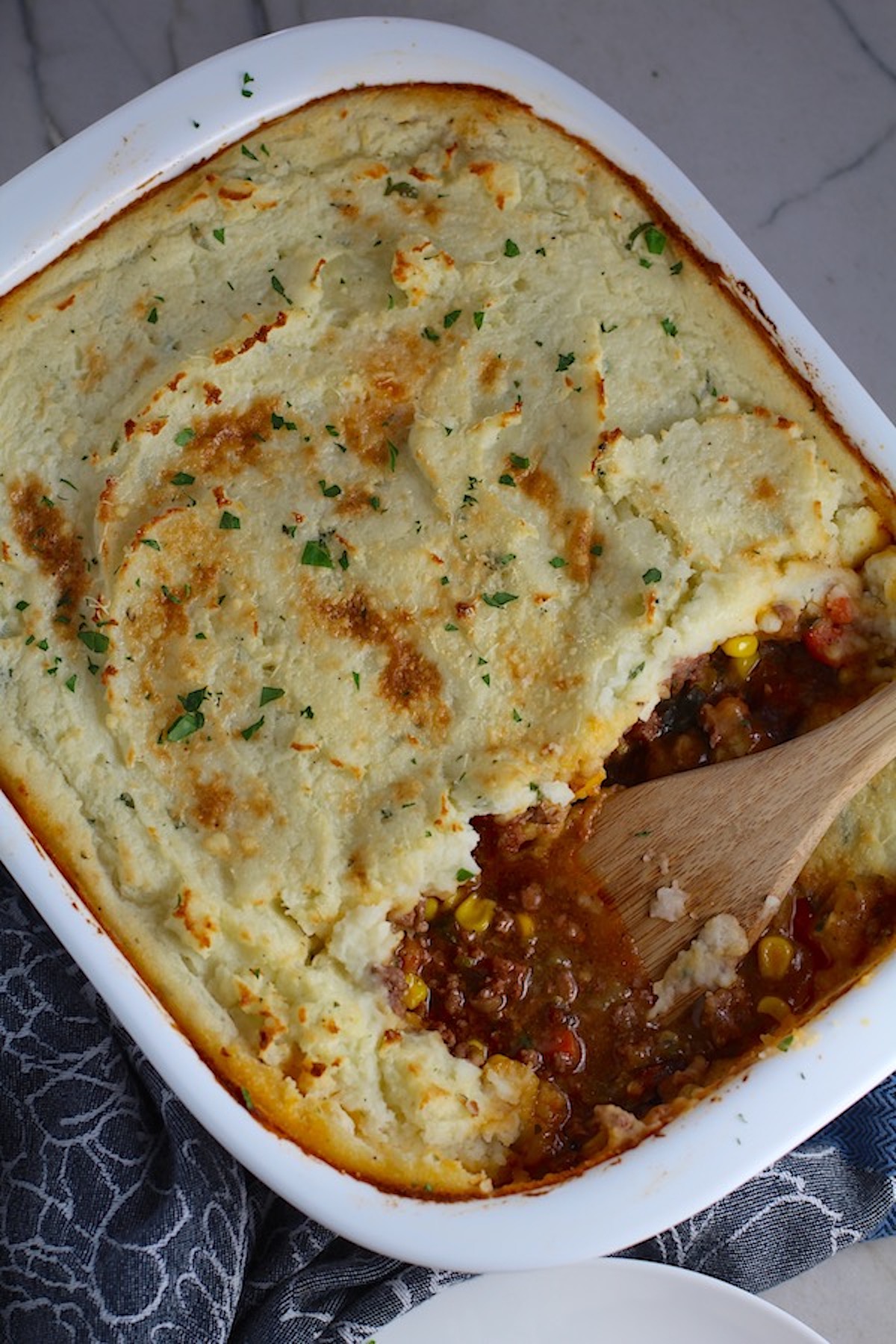 Spatula in Sausage Shepherds Pie in casserole dish. This recipe has Spanish Chorizo and ground beef cooked in a rich and savory gravy with veggies and herbs.  Creamy mashed potatoes sit on top with manchego, parmesan, and garlic.