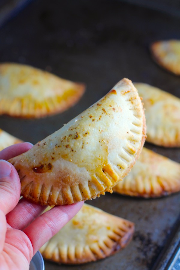 Hand holding a Ground Beef Empanada with more on a pan. Flaky, buttery pastry on the outside with a savory, smokey, salty ground beef filling.