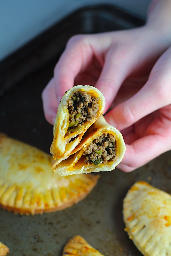Hands holding a Ground Beef Empanada with more on a pan. Flaky, buttery pastry on the outside with a savory, smokey, salty ground beef filling.