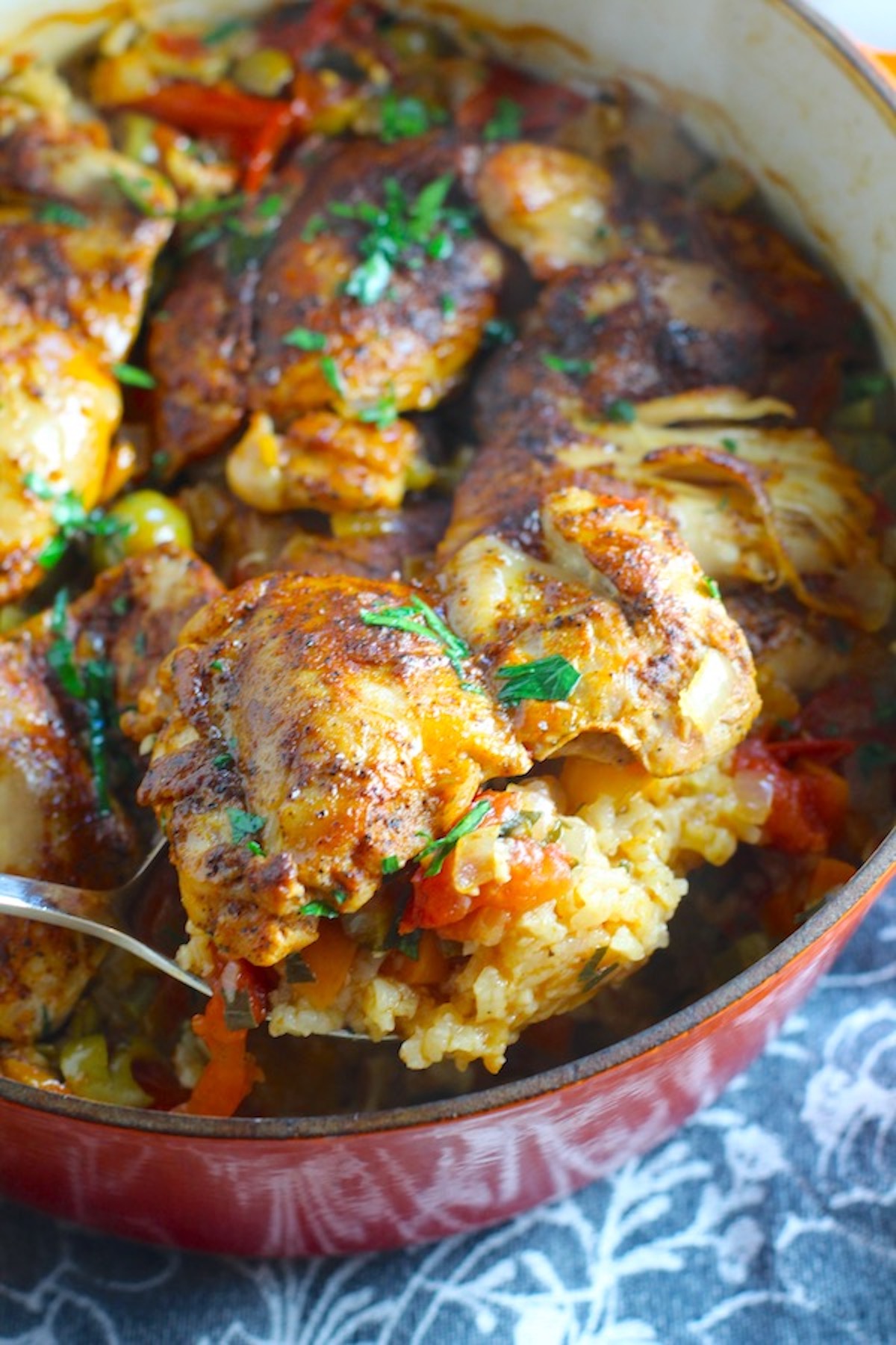 Spoon scooping Spanish Chicken and Rice up with olives and veggies from a pot on a blue towel. 