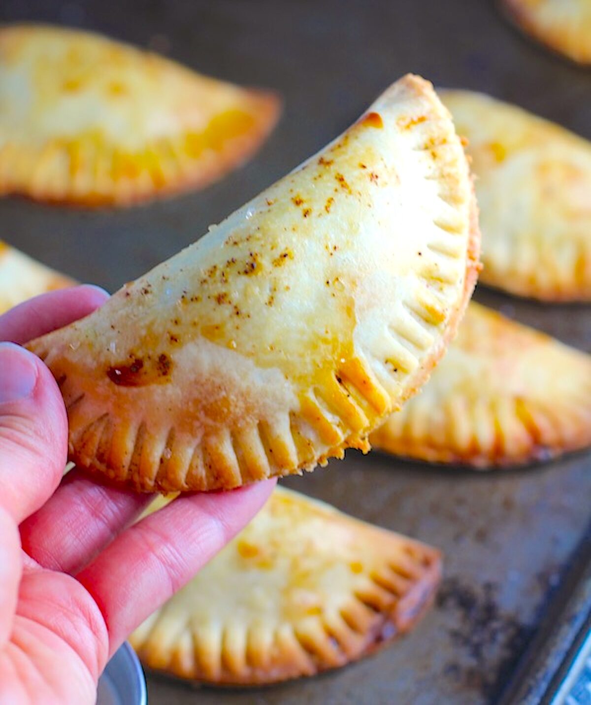 Hand holding one of the baked spiced beef empanadas over the pan of the other empanadas.